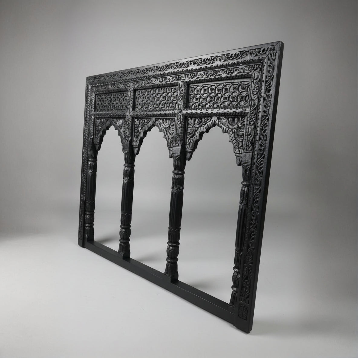 The Jharokha Wall Hanging is a carved, rectangular black wooden frame featuring intricate patterns and three arched openings that echo traditional architectural window designs. Displayed against a plain grey background, this exquisite piece of wooden décor beautifully showcases expert handicraft.