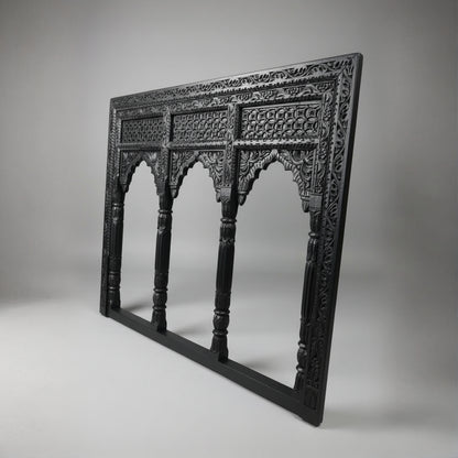 The Jharokha Wall Hanging is a carved, rectangular black wooden frame featuring intricate patterns and three arched openings that echo traditional architectural window designs. Displayed against a plain grey background, this exquisite piece of wooden décor beautifully showcases expert handicraft.