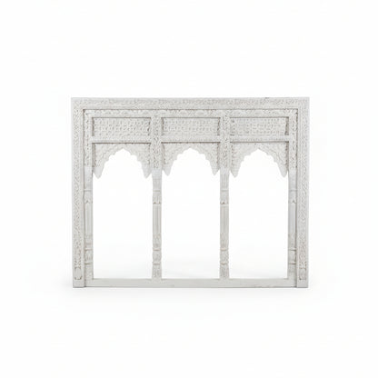 The Jharokha Wall Hanging is an ornate white carved wooden frame featuring three arches with intricate patterns and detailed columns, inspired by traditional architectural designs. Set against a plain white background, this stunning piece of handicraft is perfect for adding sophisticated decor to any space.