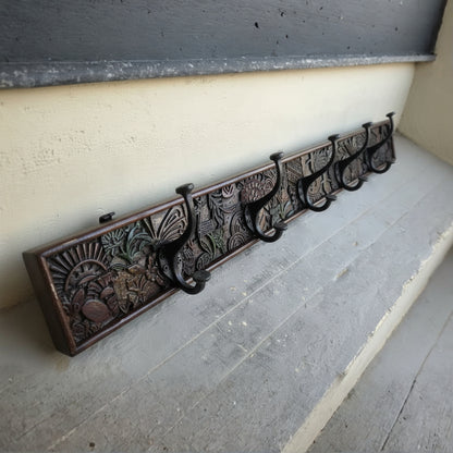 Wooden wall-mounted coat rack featuring intricate tribal hand-carved patterns with multiple black metal hooks, combining traditional decor with functional design.