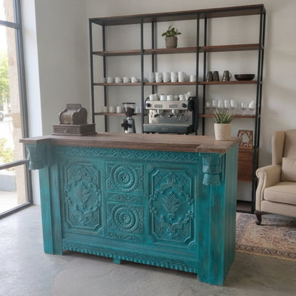 Handcrafted wooden service counter with intricate tribal-inspired carvings in a teal finish, set in a modern space with open shelving and coffee equipment.