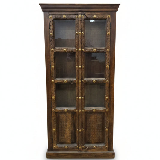 Tall wooden display cabinet with glass-paneled doors, featuring intricate tribal-style carvings and decorative brass accents, combining rustic charm and durable craftsmanship.
