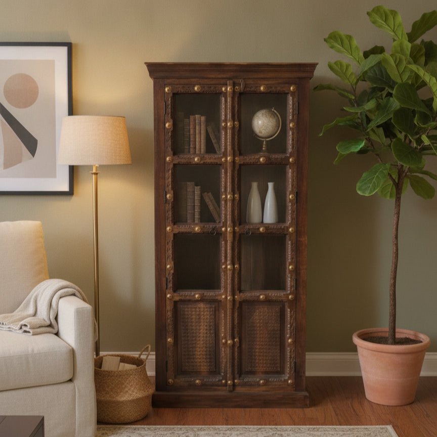 Solid wood display cabinet with intricate tribal-style handcrafted detailing, showcasing books, decorative vases, and a globe, set in a warm, cozy living room decor.